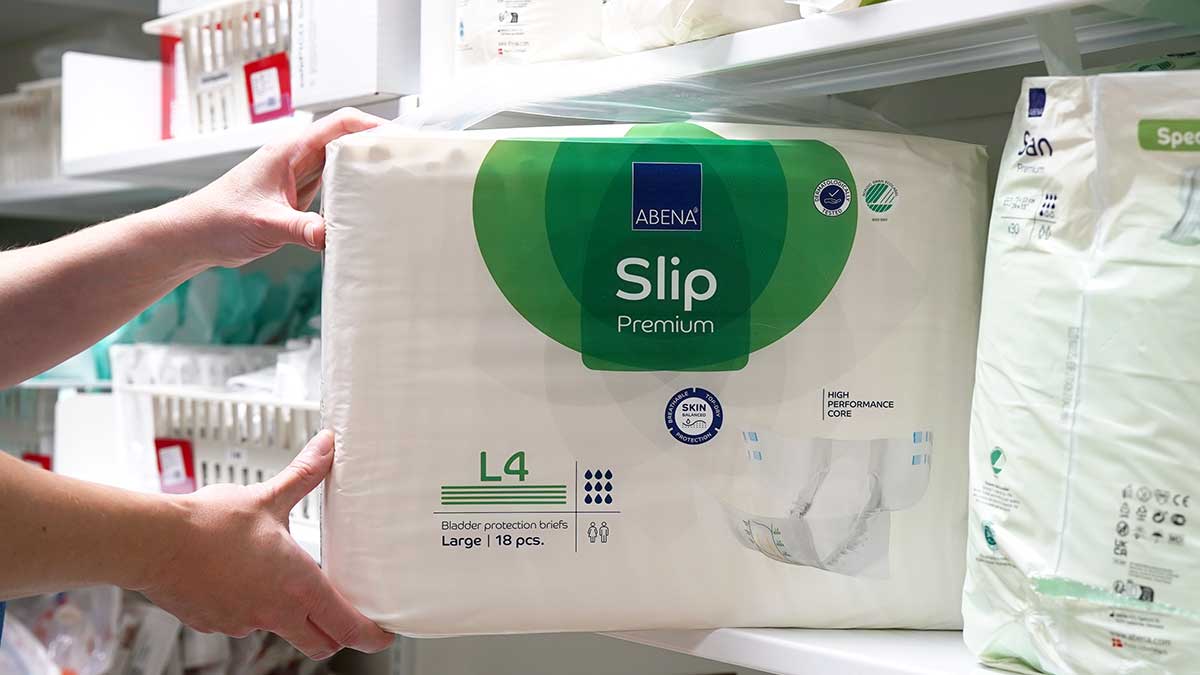 Hands placing ABENA Slip Premium incontinence product with ecolabels on a shelf